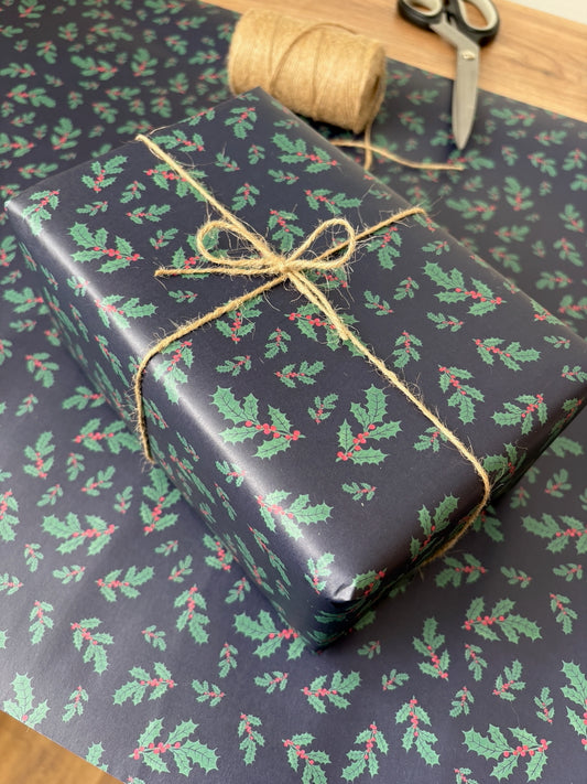 A wrapped gift box covered in dark navy holiday wrapping paper with a green holly and red berry print, tied with natural twine. Matching wrapping paper is spread underneath, with a spool of twine and scissors visible on a wooden surface.