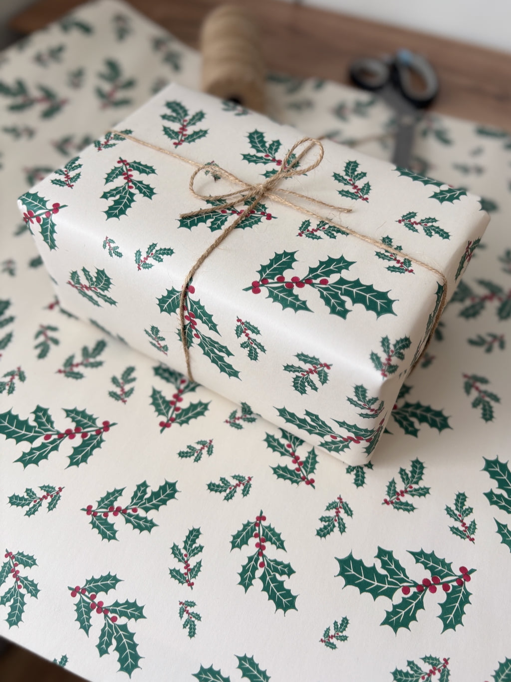 A wrapped gift box tied with twine sits on matching holiday wrapping paper printed with holly leaves and red berries. A spool of twine and scissors are visible in the background.