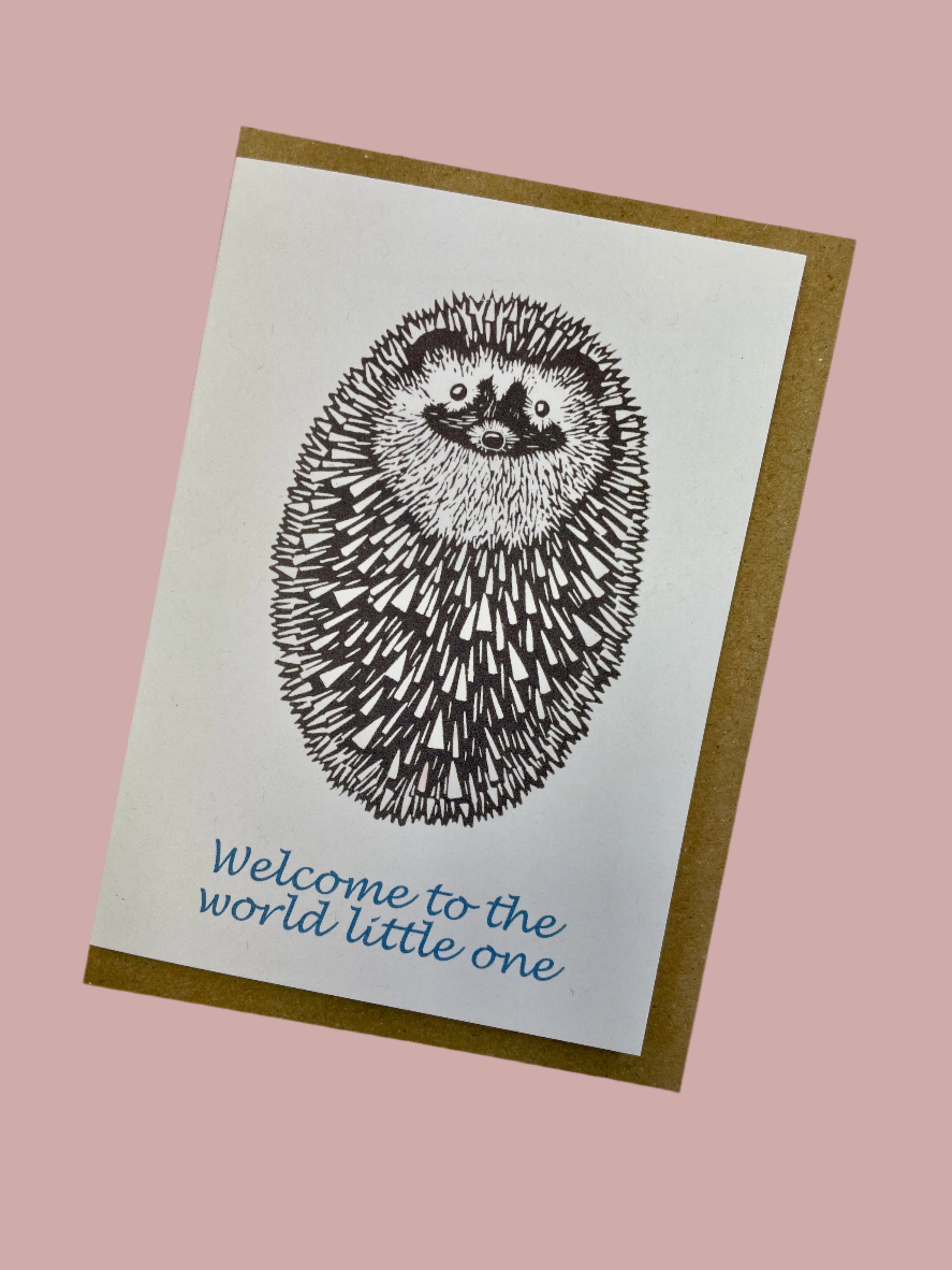 White A6 new baby card featuring a curled oval ball lino print hedgehog in black. With text below in blue 'Welcome to the world little one', with brown envelope. Card on a baby pink background.