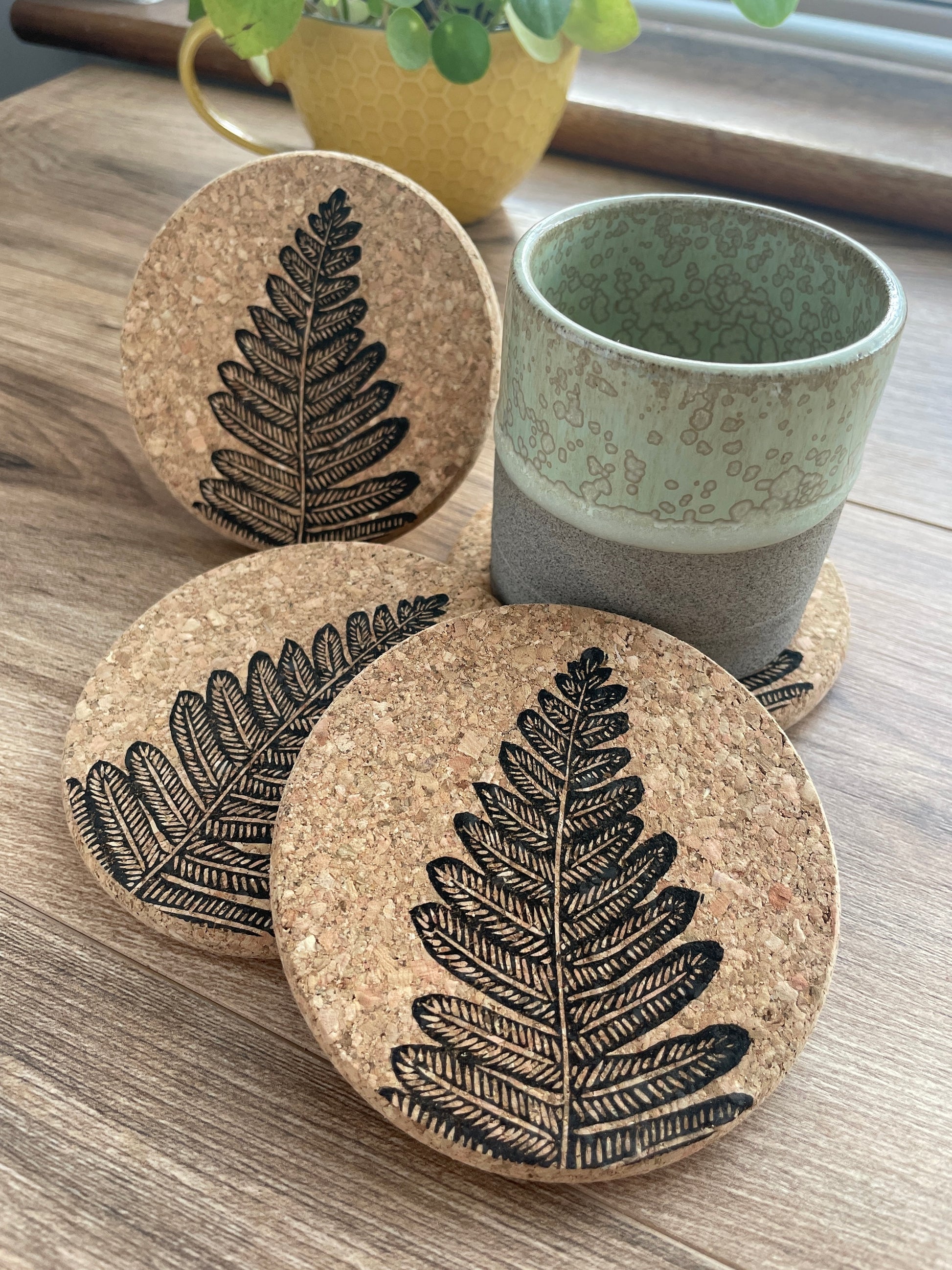 Cork coasters featuring black fern linocut print, 3 upright and one under a green pottery mug with yellow plant pot in background
