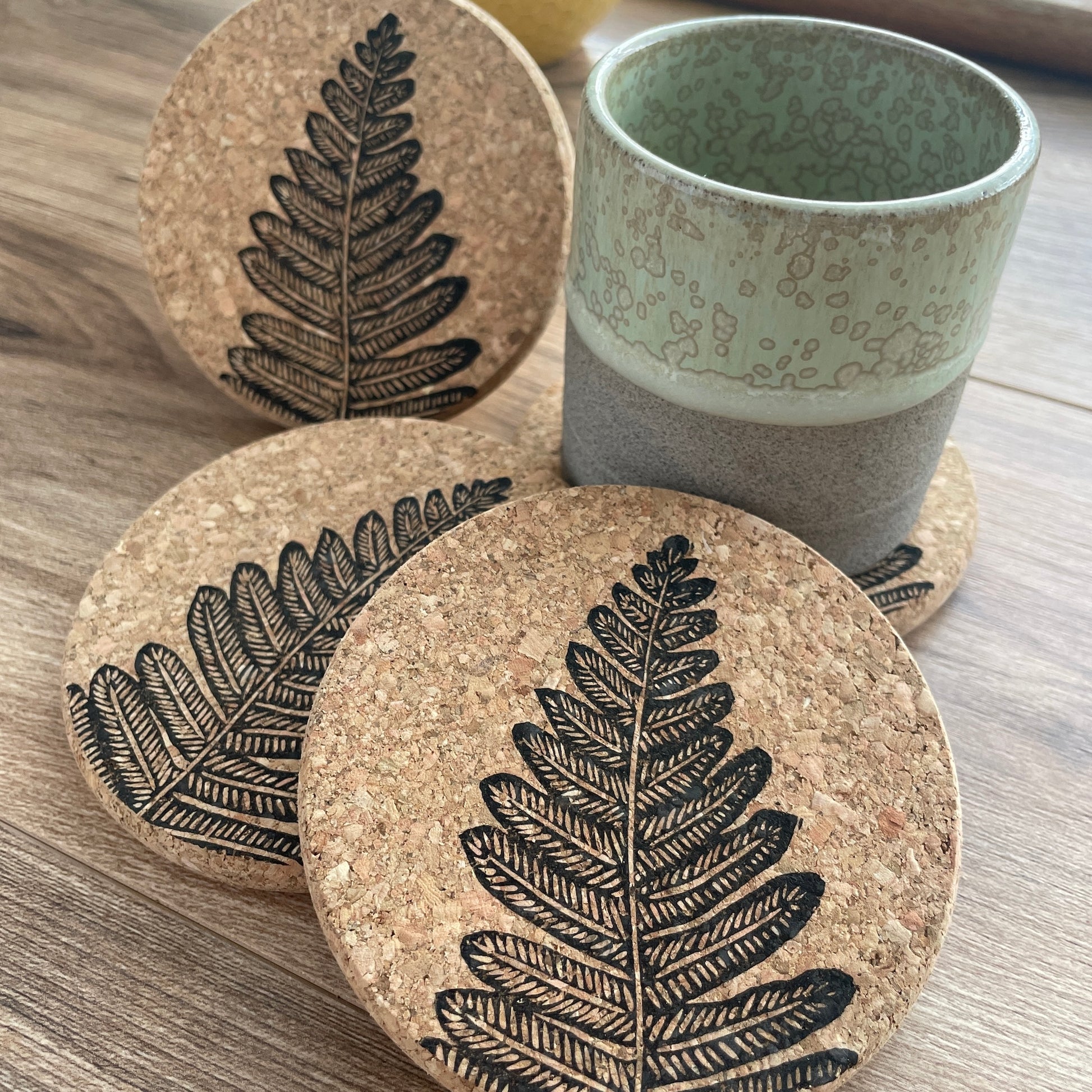Cork coasters featuring black fern linocut print, 3 upright and one under a green pottery mug