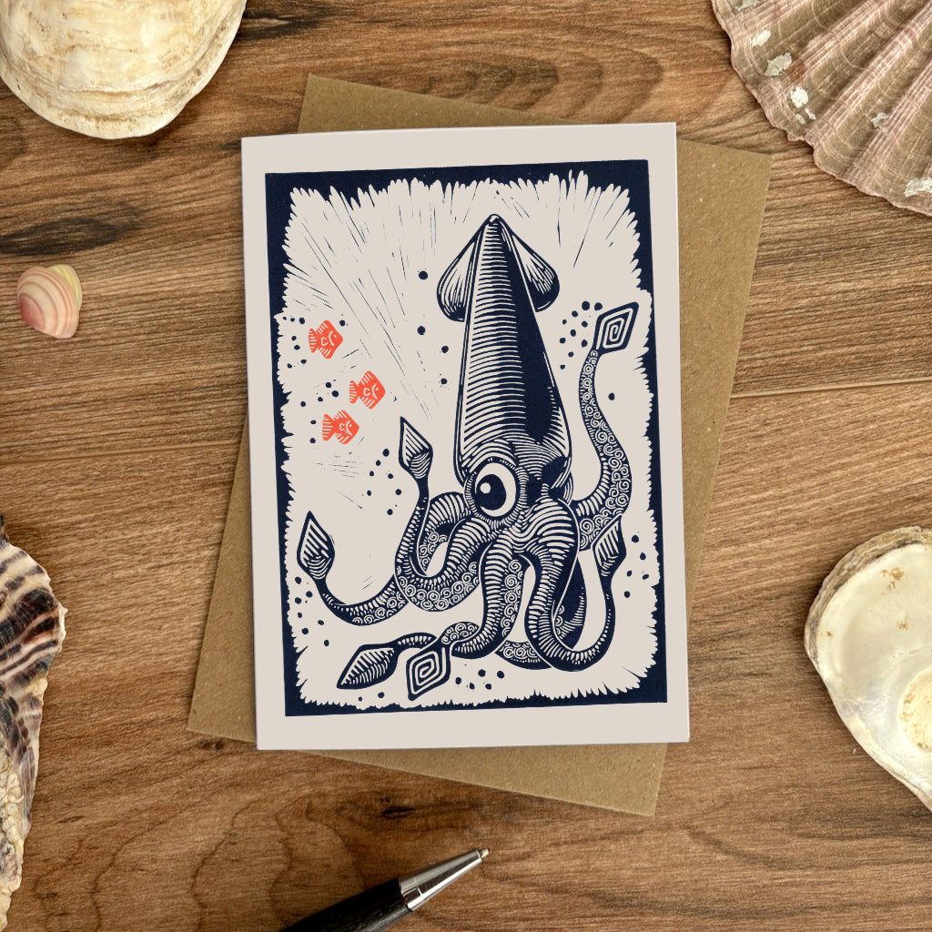 A card featuring a navy blue ink linocut design of a squid on a white background, placed on top of a brown kraft envelope over a wooden surface with seashells around it.