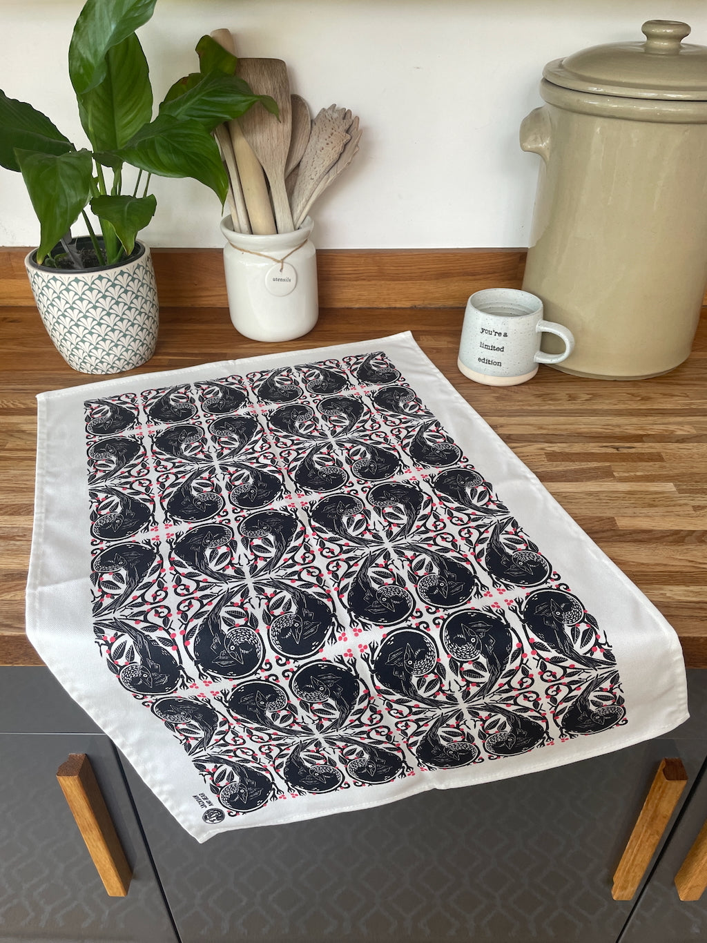 Tea towel with jackdaw repeat pattern in red, black and white pattern on a wooden worktop with kitchen items.