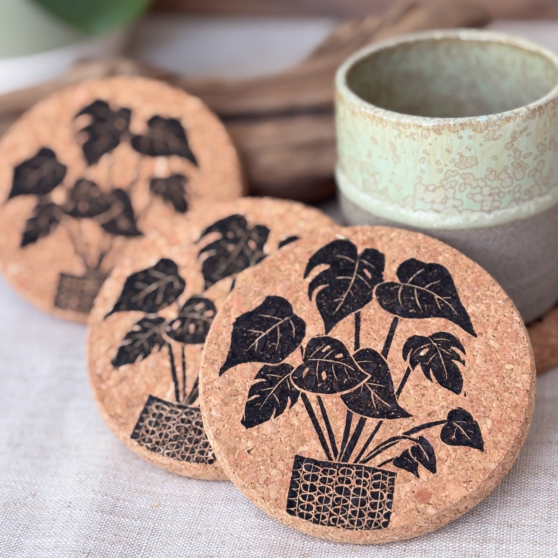 Cork coasters with Monstera house plant design next to a ceramic cup on a light fabric background