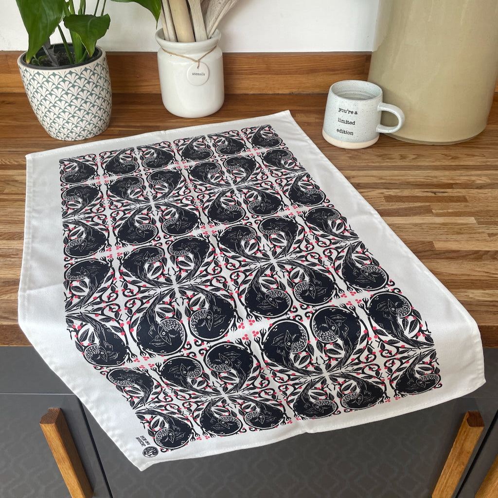 A red and black geometric patterned tea towel laid out on a kitchen counter with a plant, wooden spoons, a mug, and a crockery jar in the background.