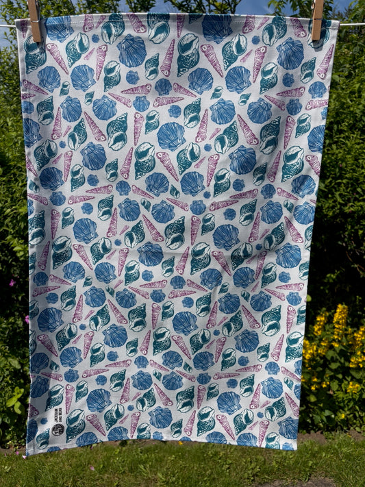 Sea shell linocut tea towel pegged on washing line with a garden hedge and wildflowers in the background