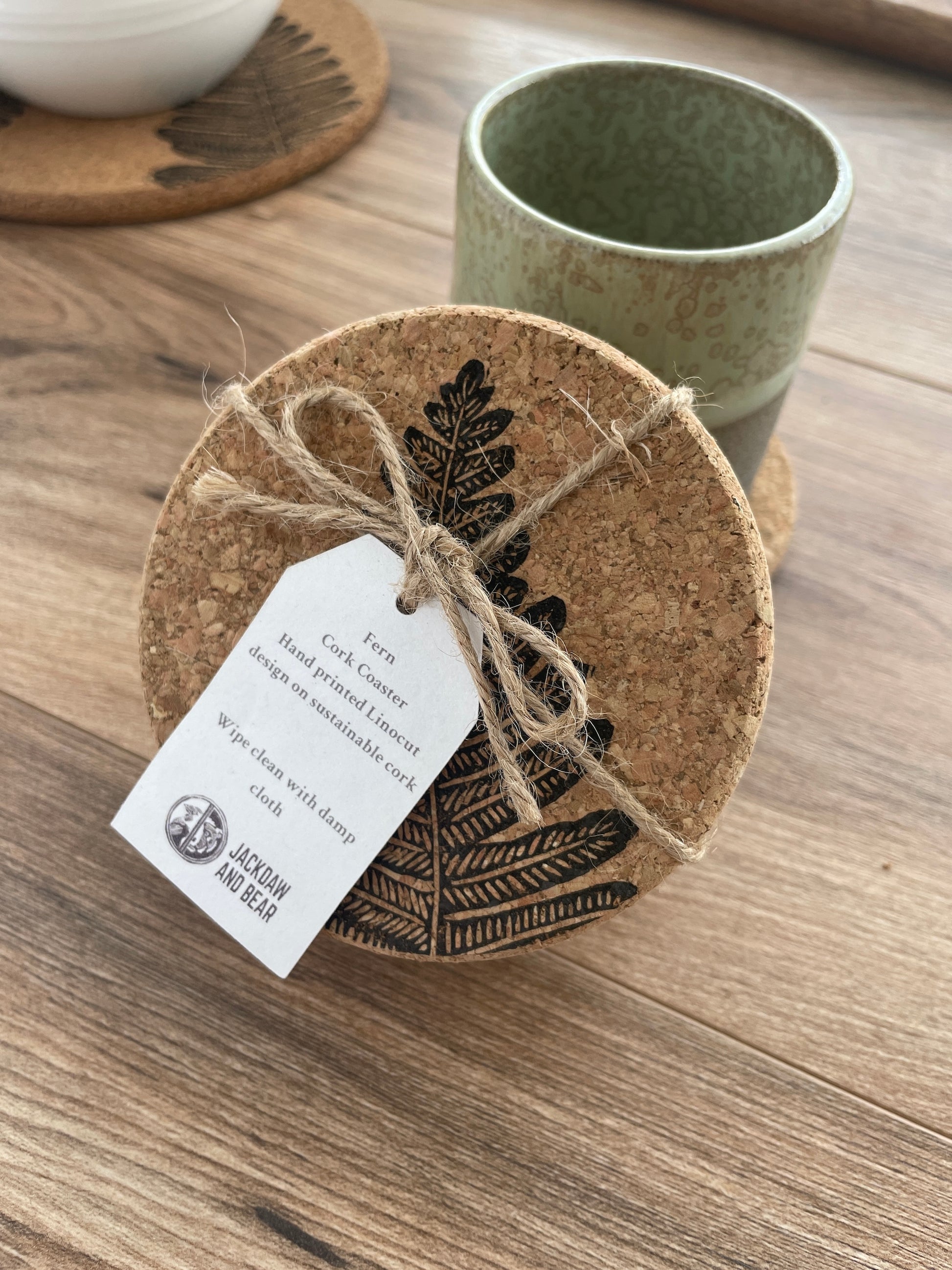 Cork coasters featuring black fern linocut print, set of 4 packaged with brown twine and into tag leaning against a green pottery mug with bowl on matching placemat in background