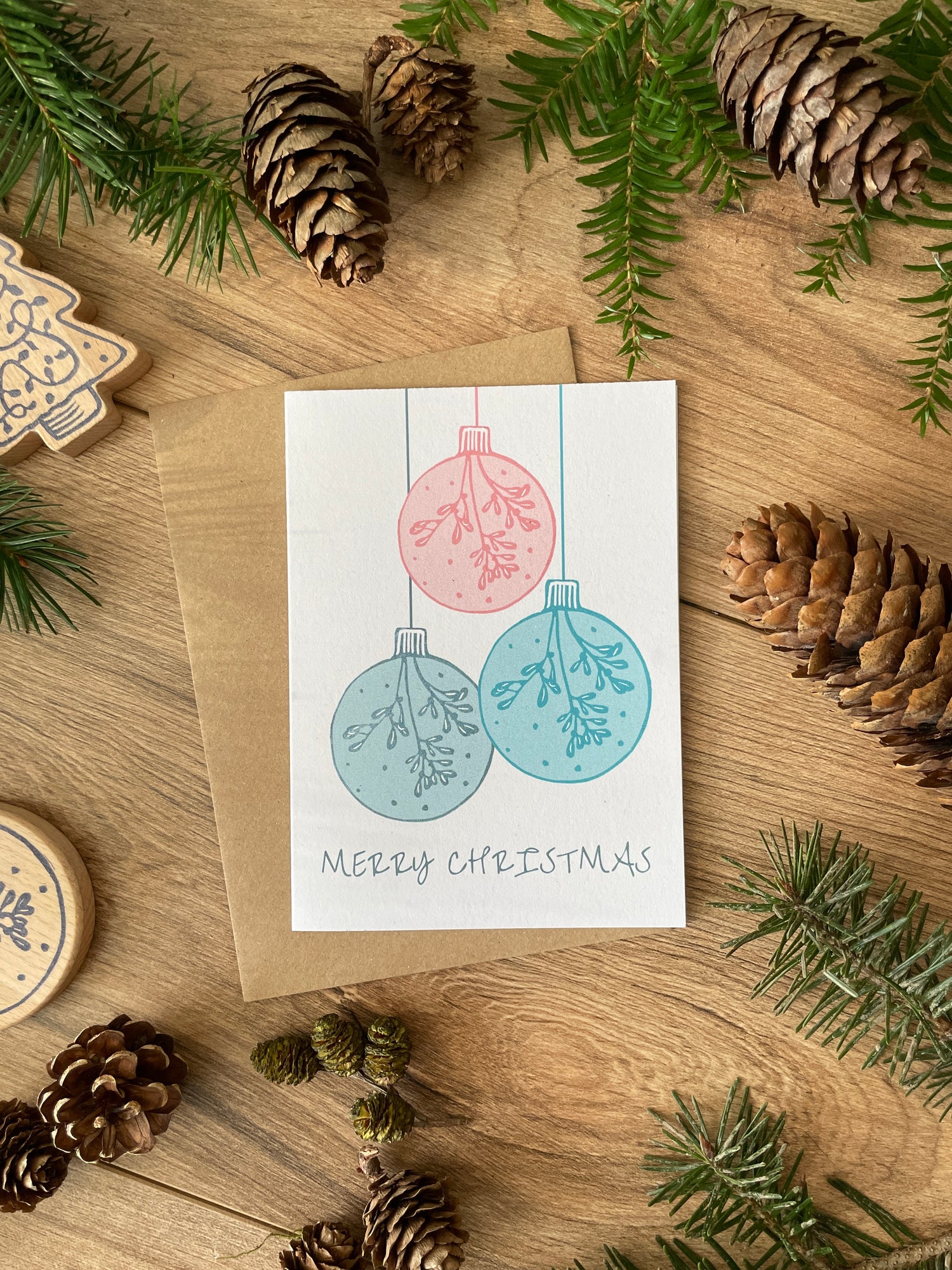 Flat lay of Christmas card featuring three baubles from linocut design in pink, blue and navy on background of fir and pinecones