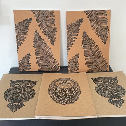 5 notebooks with linocut prints- 2 propped up with 4 fern leaf prints, and 3 flat laying with middle book featuring hedgehog print, and the two either side with an owl print.