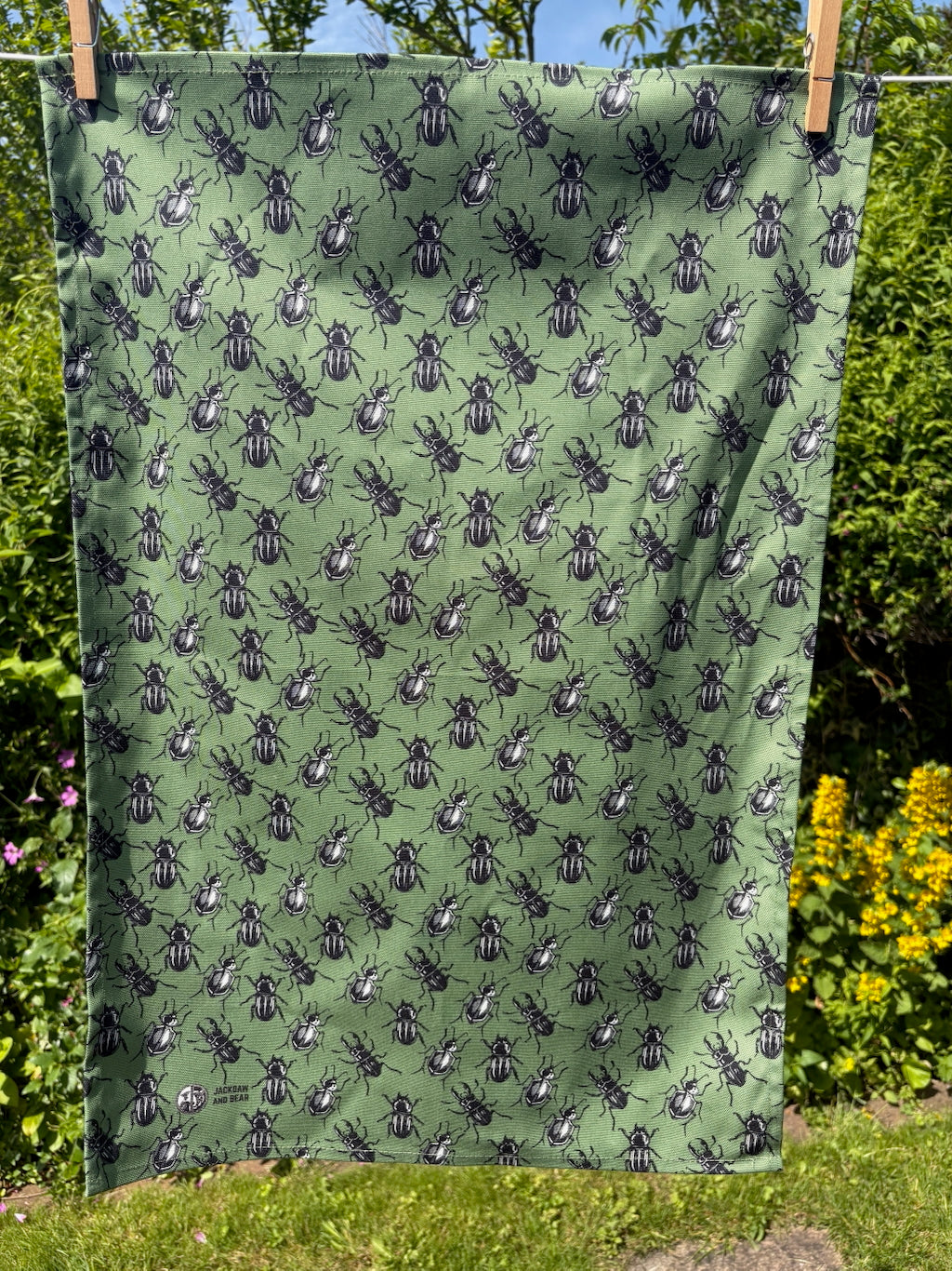 Green beetle linocut tea towel pegged on a washing line with a garden hedge and wildflowers in the background
