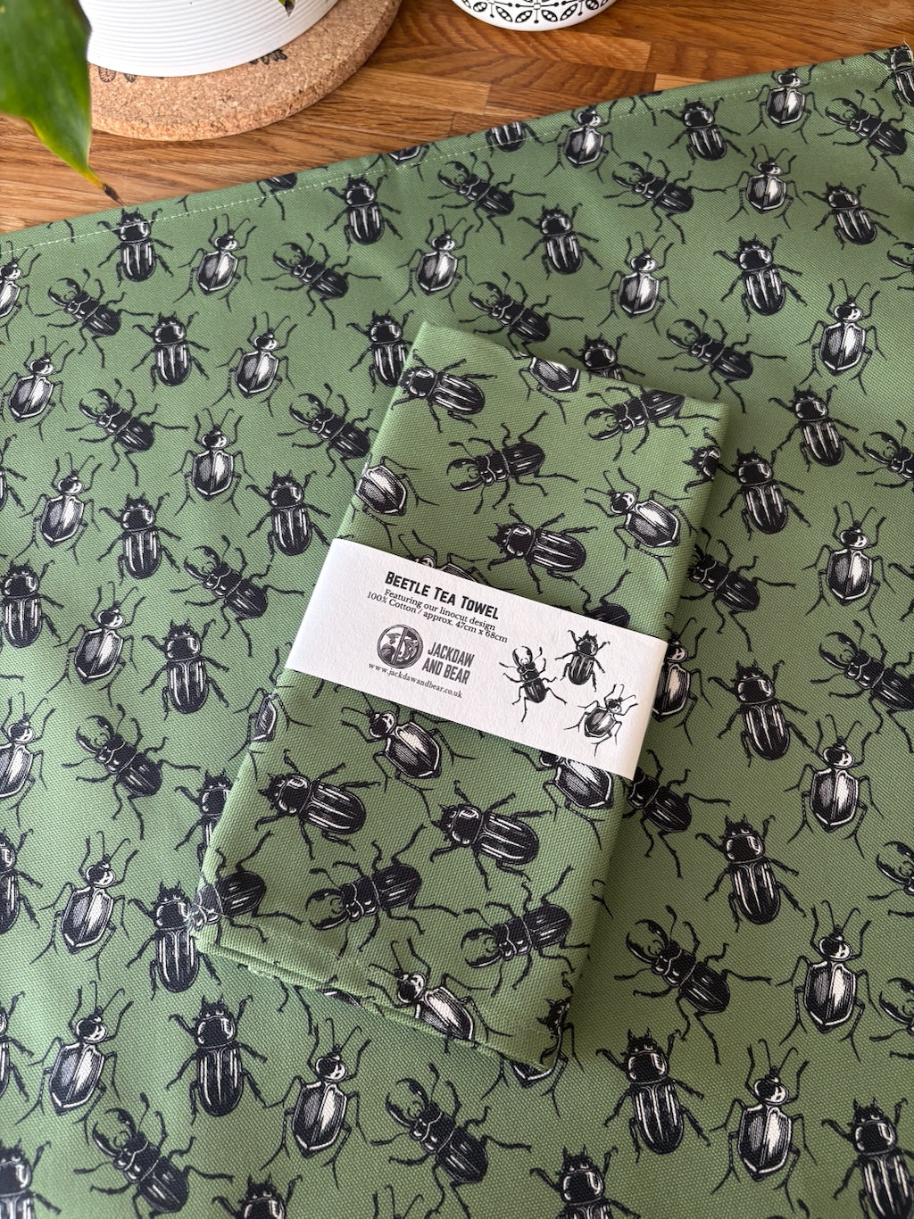 Folded green tea towel with beetle pattern in recycled card belly band packaging, laid on top of the same green beetle tea towel flat on a wooden worktop
