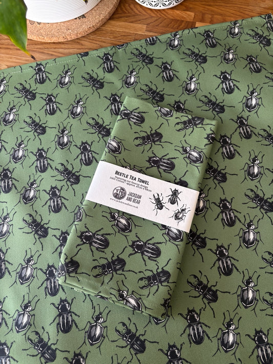 Folded green tea towel with beetle pattern in recycled card belly band packaging, laid on top of the same green beetle tea towel flat on a wooden worktop
