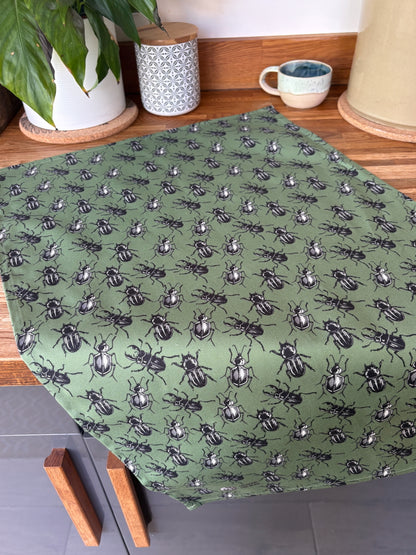 Green beetle patterned linocut print tea towel laid flat on wooden worktop, styled with a houseplant and cup against grey kitchen cupboards
