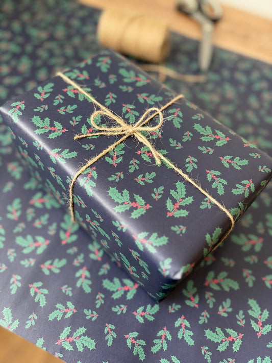 A wrapped gift box with navy blue wrapping paper featuring a green holly and red berry pattern, tied with natural jute twine, placed on matching wrapping paper.