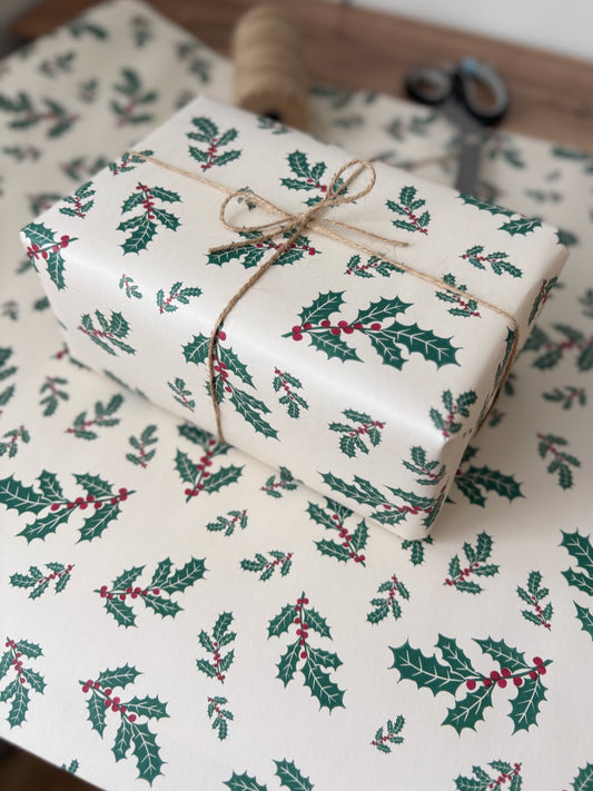 A wrapped gift box tied with twine sits on matching holiday wrapping paper printed with holly leaves and red berries. A spool of twine and scissors are visible in the background.