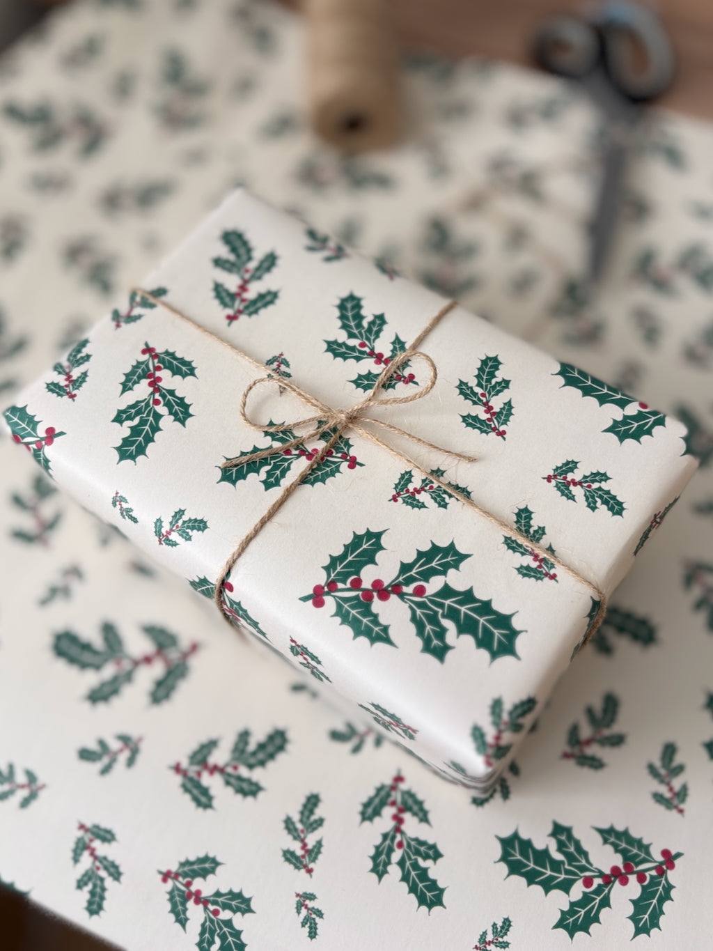 A wrapped gift box covered in cream wrapping paper with a green holly and red berry print, tied with a natural twine bow. Matching wrapping paper is spread out underneath, with a blurred background showing crafting tools.