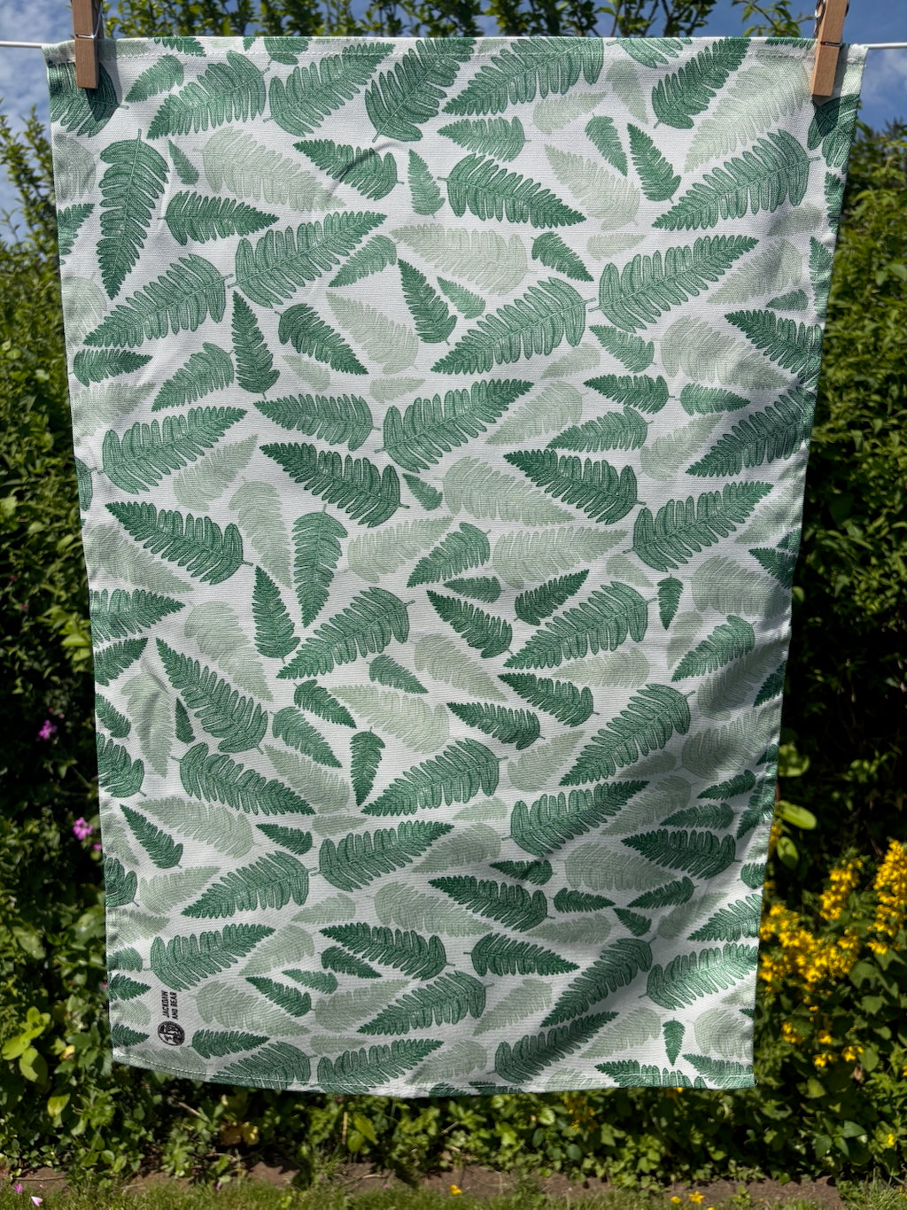Green fern linocut tea towel pegged on a washing line with a garden hedge and wildflowers in the background