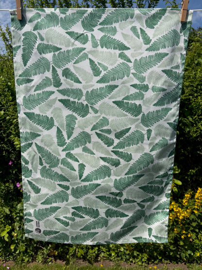 Green fern linocut tea towel pegged on a washing line with a garden hedge and wildflowers in the background