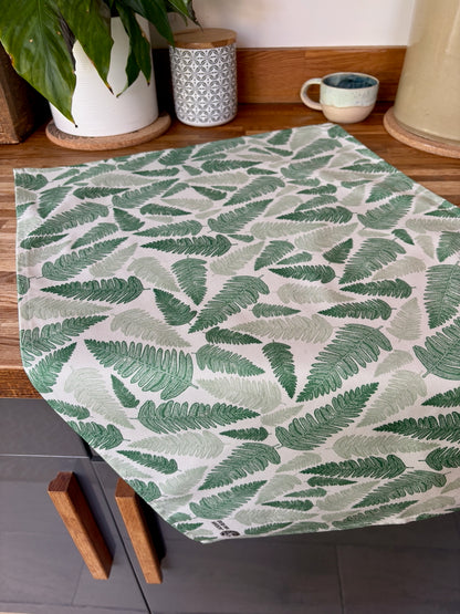 Green fern linocut print tea towel laid flat on wooden worktop, styled with a houseplant and cup against grey kitchen cupboards