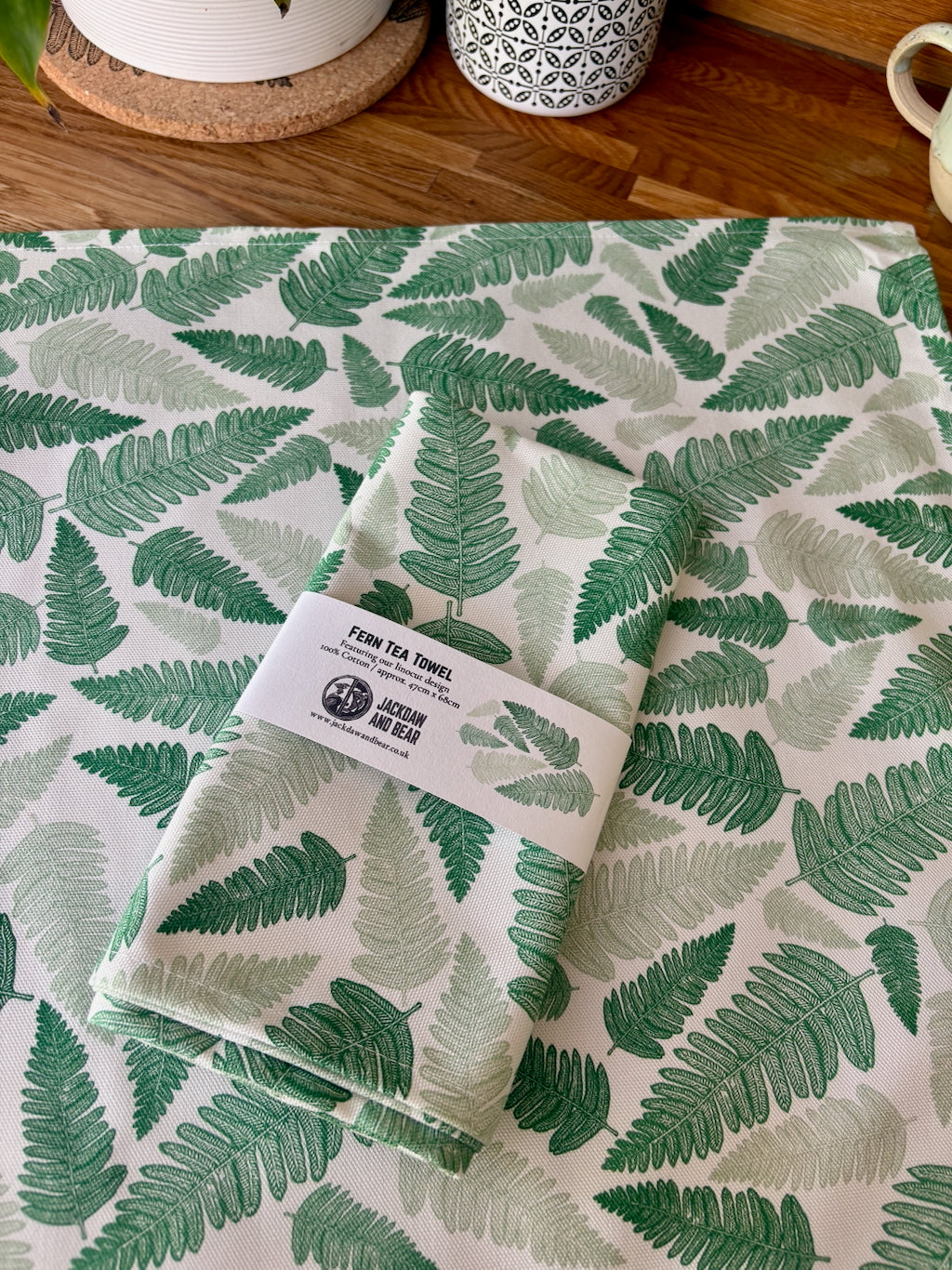 Folded Fern tea towel in recycled card belly band  packaging, laid on top of the same green fern tea towel flat on a wooden worktop
