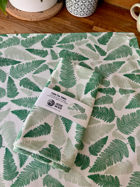 Folded Fern tea towel in recycled card belly band  packaging, laid on top of the same green fern tea towel flat on a wooden worktop