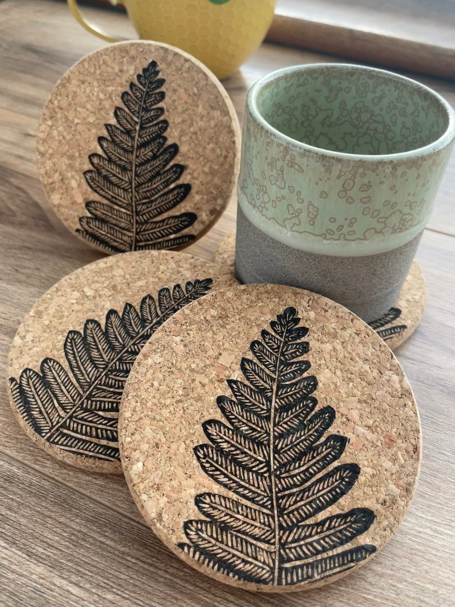 Cork coasters featuring black fern linocut print, 3 upright and one under a green pottery mug with yellow plant pot in background