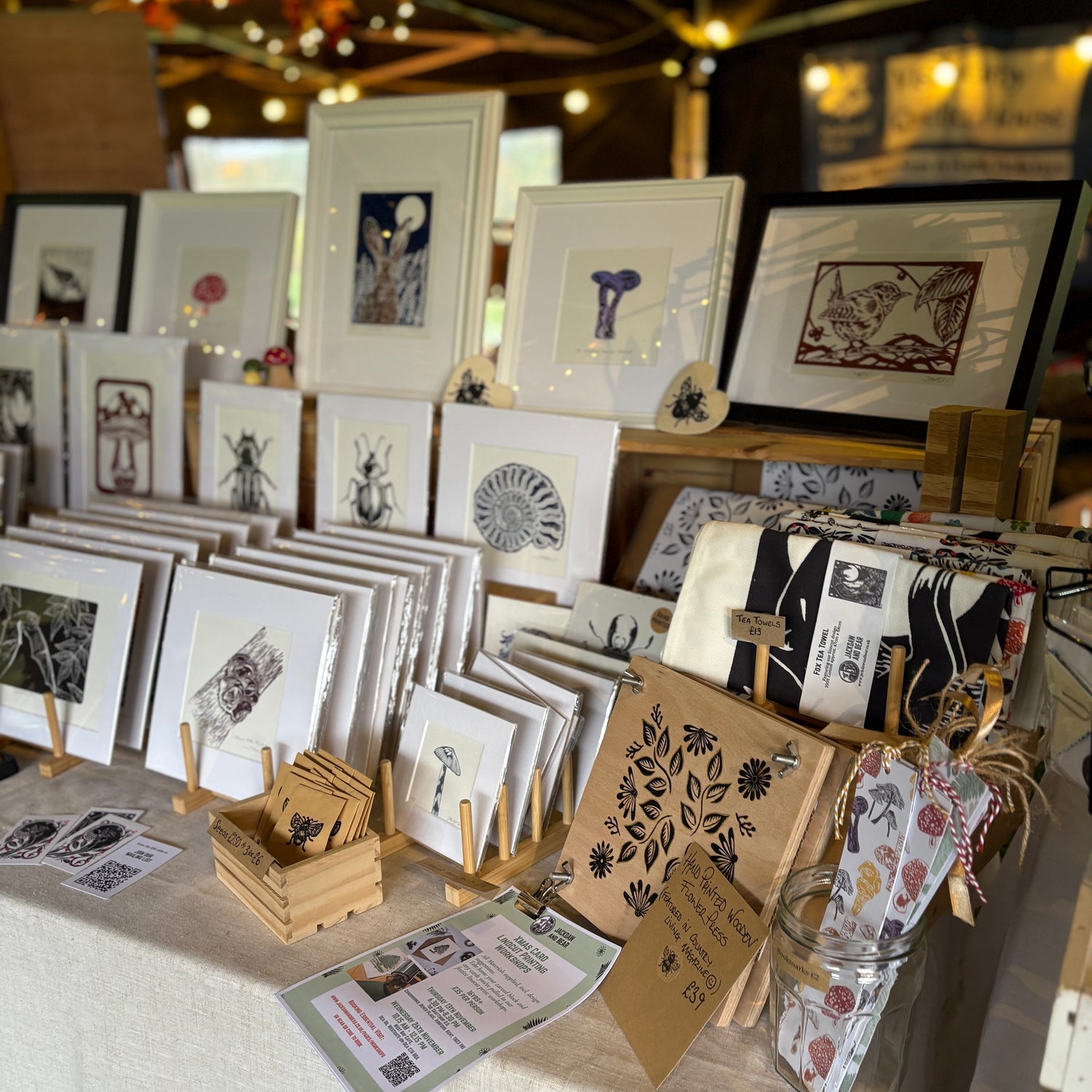 A market stall display featuring various framed art prints, small wooden blocks, and patterned paper tags on a white tablecloth, with warm string lights in the background.