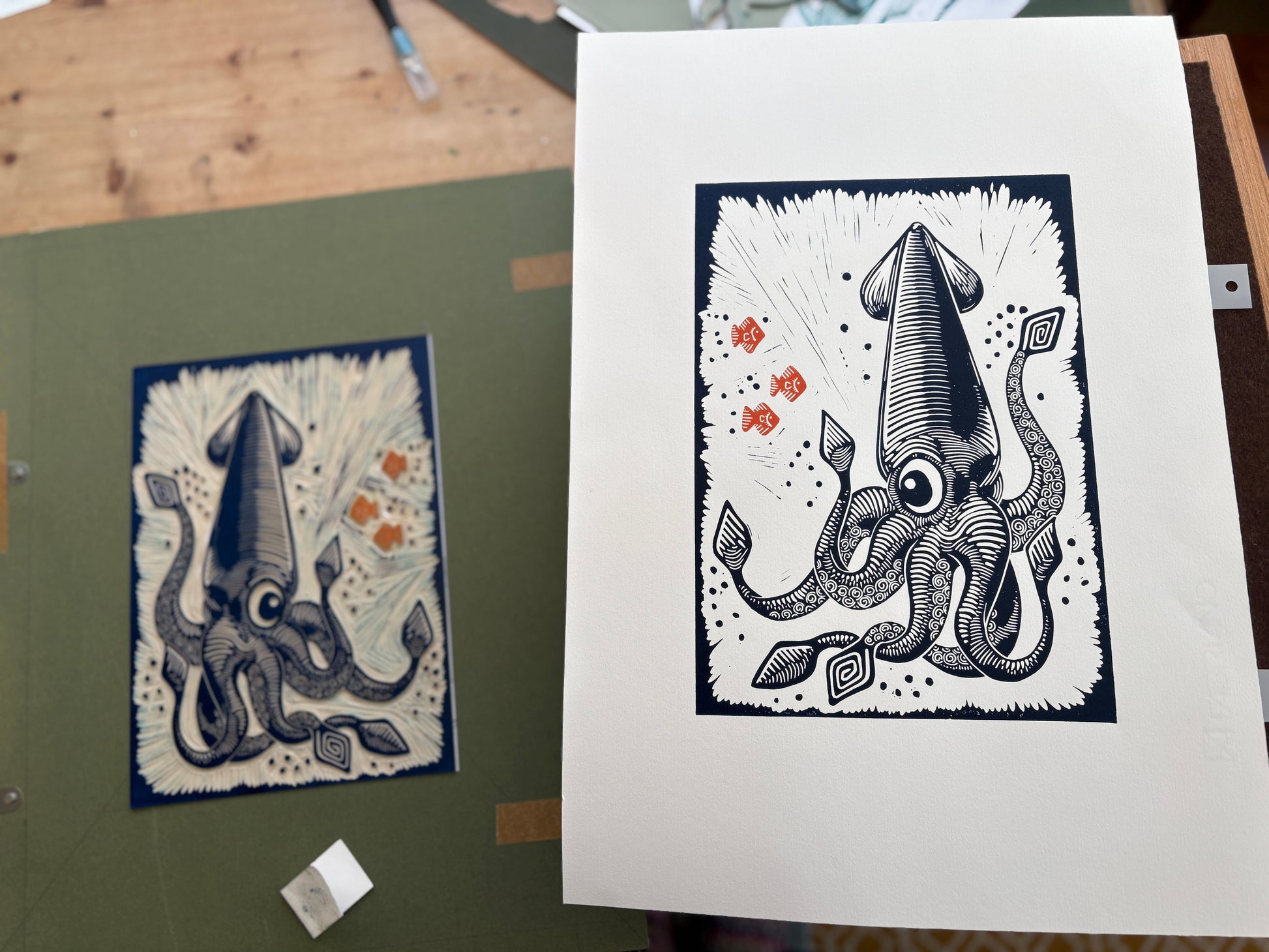 Linocut print and block of a squid design on paper with a green surface and wooden desk in the background.