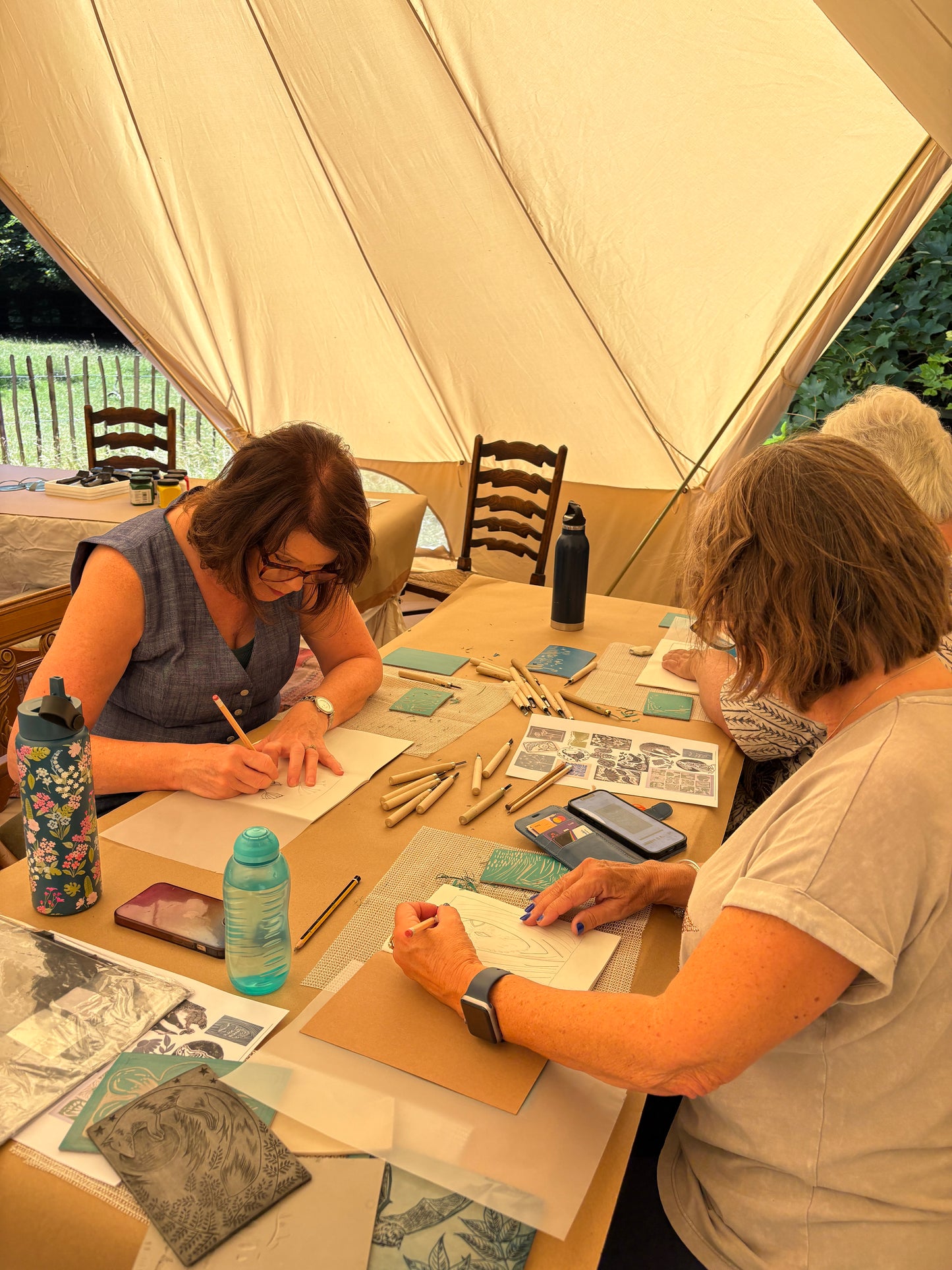 Three women sitting at a wooden table inside a large cream-colored canvas tent, working on handmade prints with pencils and tools. Various art supplies, water bottles, and papers are spread across the table.