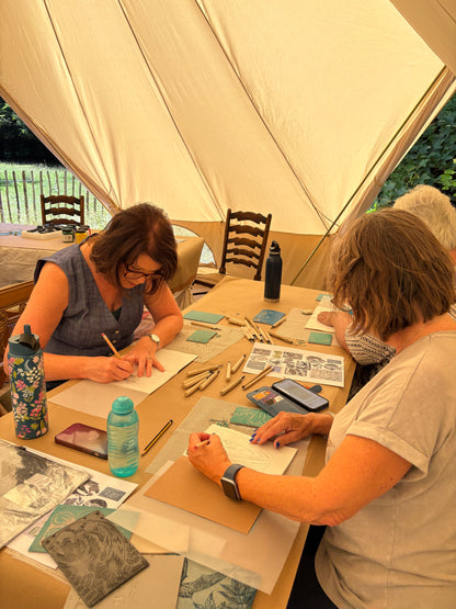 Three women sitting at a wooden table inside a large cream-colored canvas tent, working on handmade prints with pencils and tools. Various art supplies, water bottles, and papers are spread across the table.