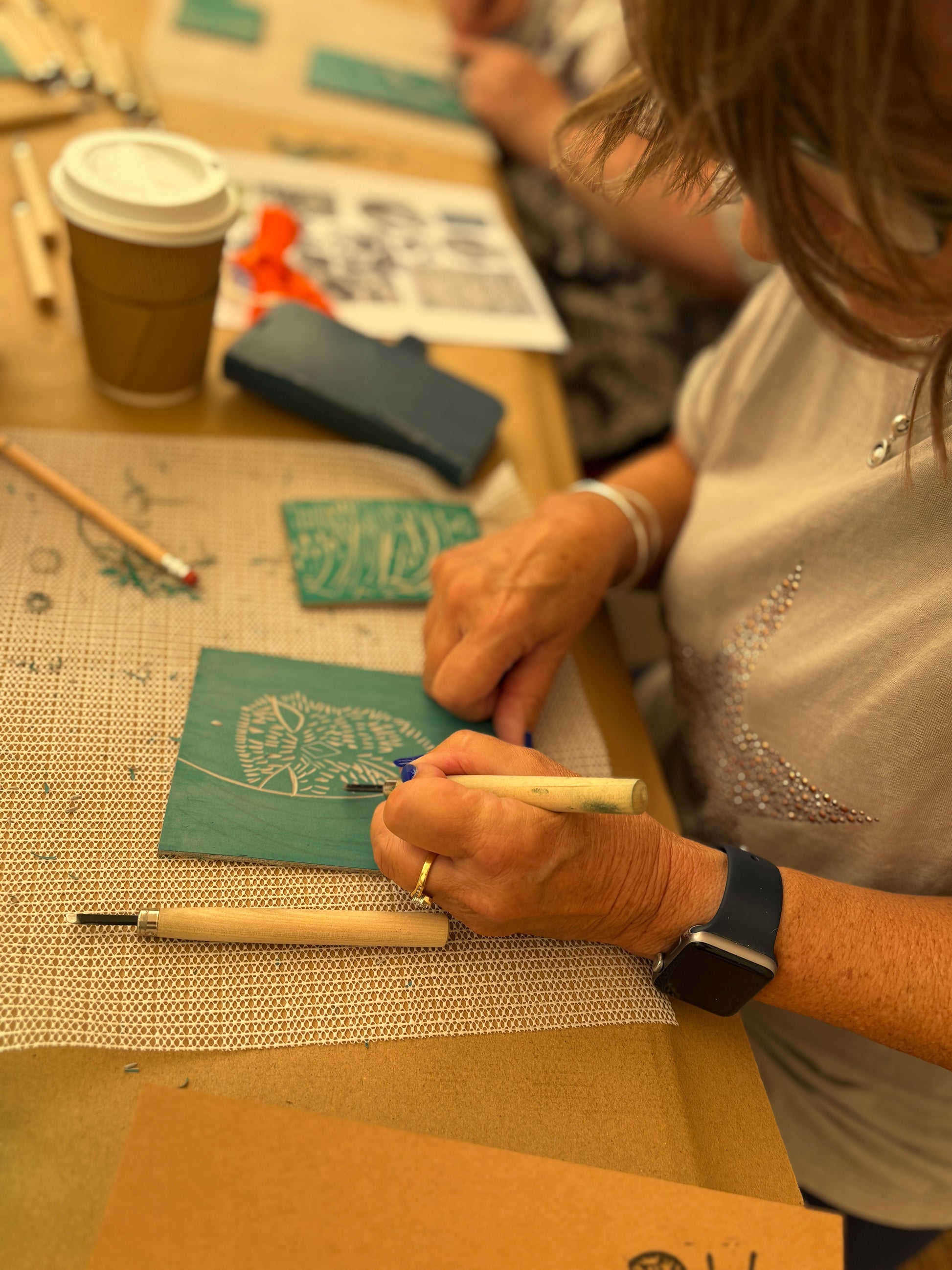 A close-up lifestyle shot of a woman carving an oak tree block print on a green wood panel at a workshop table. She is holding a carving tool over a textured mat, with other crafting supplies, a coffee cup, and another person blurred in the background.