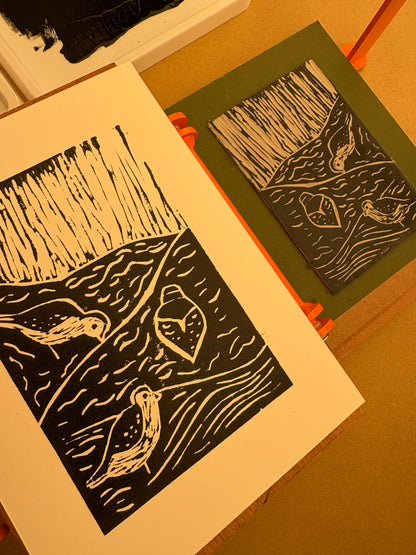 A close-up of a black and cream linocut  print featuring birds and abstract background, shown on a white sheet of paper. A block in a green registration sheet is visible in the background on level press.