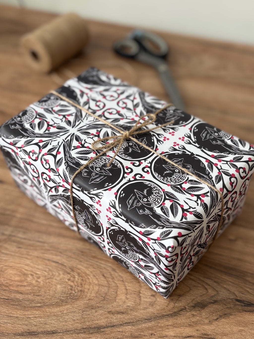A rectangular gift box wrapped in black, white, and red botanical jackdaw patterned paper, tied with twine, sitting on a wooden tabletop with a twine spool and scissors blurred in the background.