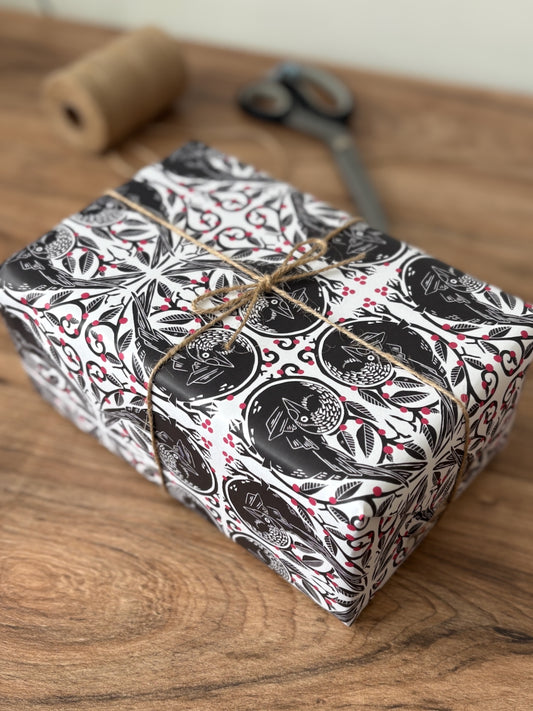 A rectangular gift box wrapped in black, white, and red botanical jackdaw patterned paper, tied with twine, sitting on a wooden tabletop with a twine spool and scissors blurred in the background.