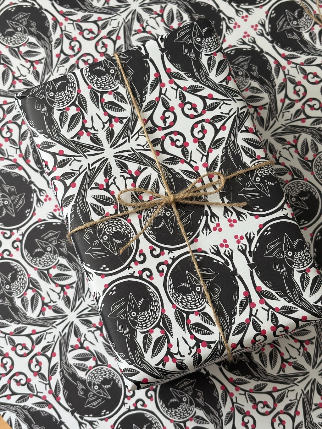 A gift box wrapped in black and white owl-patterned paper with red berries, tied with natural twine, photographed from above on a matching patterned surface.