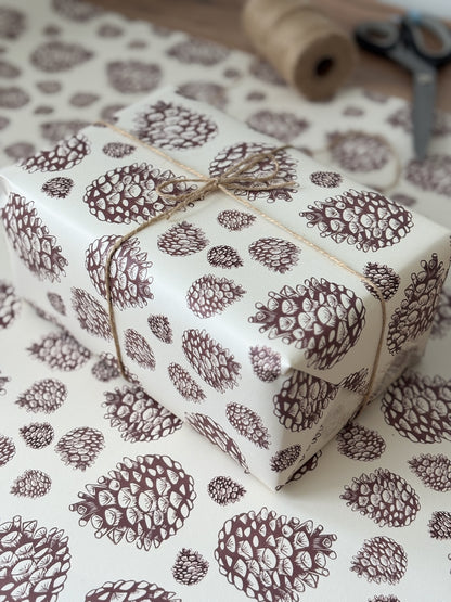 A gift-wrapped box and matching wrapping paper with a brown pinecone pattern on a white background, tied with twine, with a spool and scissors blurred in the background.