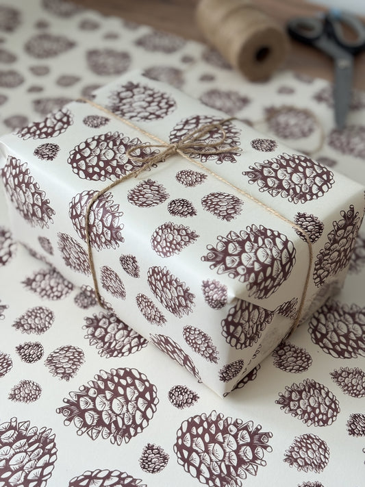 A gift-wrapped box and matching wrapping paper with a brown pinecone pattern on a white background, tied with twine, with a spool and scissors blurred in the background.