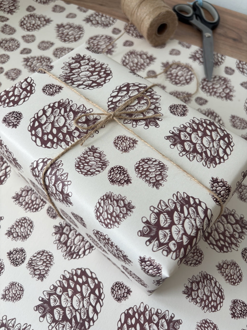 Pinecone-patterned wrapping paper on a wooden surface, with a wrapped gift box tied with twine and a spool of twine and scissors nearby.