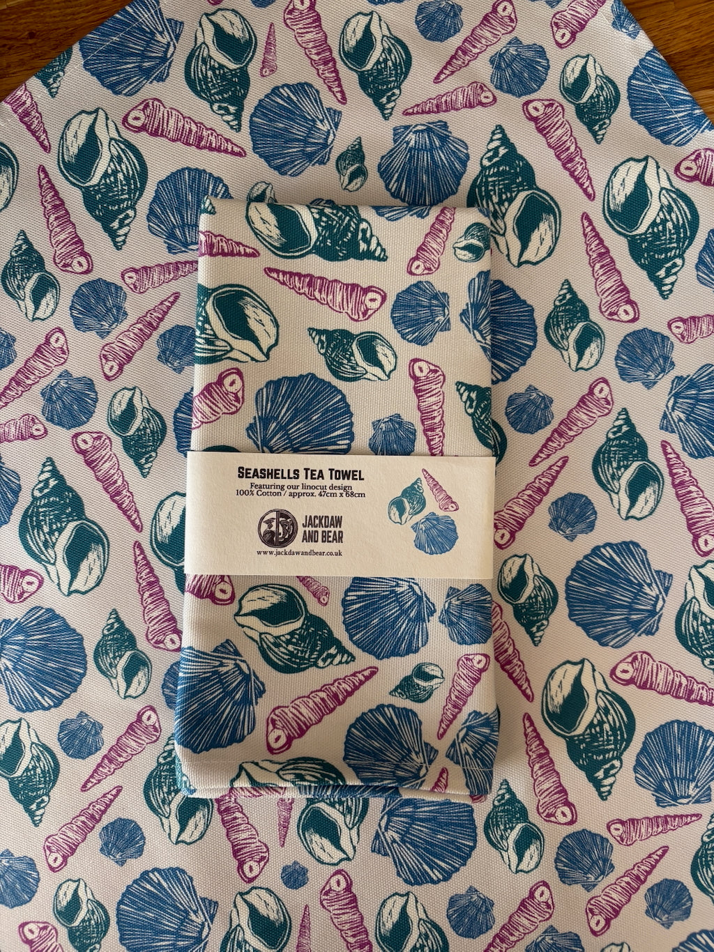 Folded sea shell tea towel in recycled card belly band packaging, laid on top of the same pink and blue shell linocut tea towel flat on a wooden worktop
