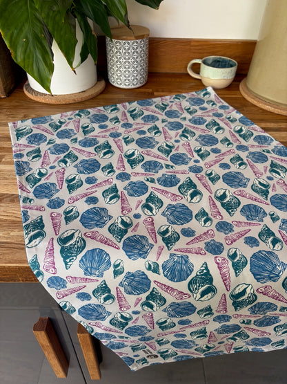 Bright sea shell linocut print tea towel laid flat on a wooden worktop, styled with a house plant and cup against grey kitchen cupboards