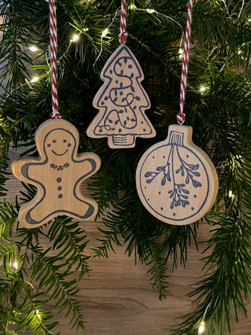 Set of 3 wooden Christmas tree decoration hanging on pine trees with fairy lights in background. Features navy linocut design of bauble, tree and gingerbread man hand printed on to beech wood paired with red and white twine