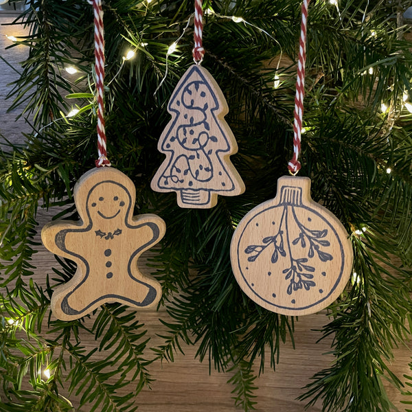 Set of 3 wooden Christmas tree decoration hanging on pine trees with fairy lights in background. Features navy linocut design of bauble, tree and gingerbread man hand printed on to beech wood paired with red and white twine